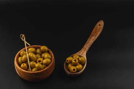 Fresh green olives are arranged in a rustic wooden bowl alongside a wooden spoon filled with more olives, set against a dark backdrop highlighting their natural color.の写真素材