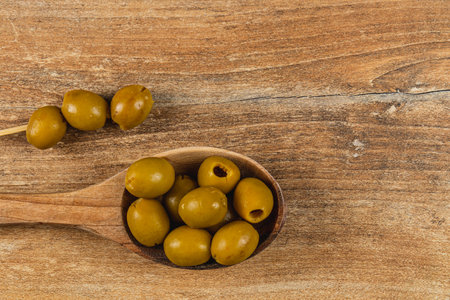 A wooden spoon holds a variety of green olives, with additional olives placed directly on the table. The rustic surface enhances the natural textures of the olives.の写真素材