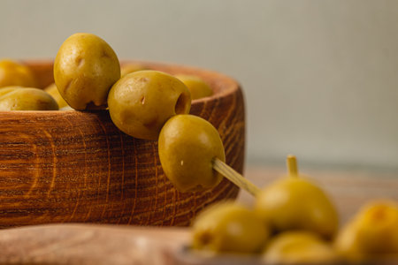 A collection of green olives arranged on a skewer rests beside a handcrafted wooden bowl filled with more olives. Natural light highlights the textures of the olives and bowl.の写真素材