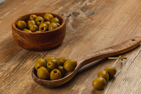 Various green olives are arranged on a wooden table, accompanied by a bowl filled with more olives. A wooden spoon rests nearby, creating an appetizing presentation.の写真素材
