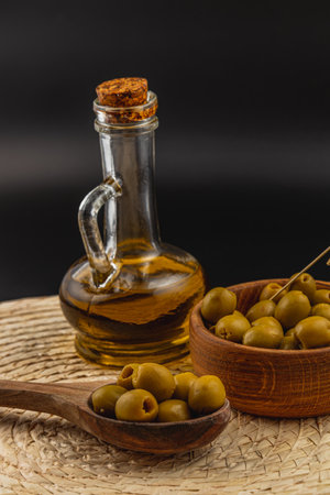 Freshly harvested green olives are placed in a wooden bowl next to a glass bottle filled with olive oil, set on a woven mat against a dark background.の写真素材