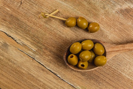 A wooden spoon holds several green olives with one olive skewered nearby. The background features a textured wooden surface, enhancing the natural look.の写真素材