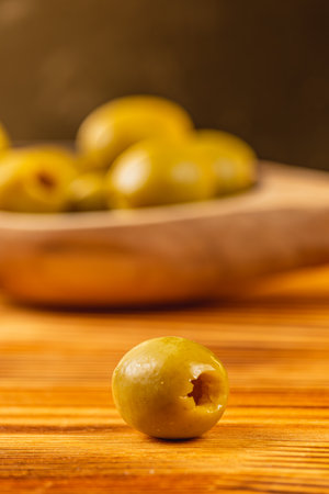 A single green olive rests on a wooden surface, with a bowl of more olives blurred in the background. This setting suggests a culinary context or snack preparation.の写真素材