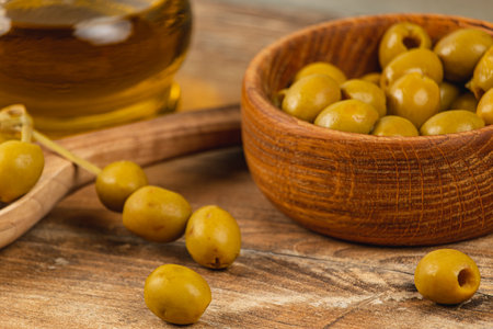 A bowl of fresh green olives sits on a wooden table alongside a bottle of olive oil. The setting features rustic textures and natural tones, evoking a Mediterranean atmosphere.の写真素材