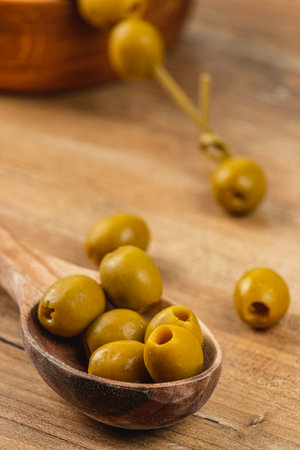 A wooden spoon holds a small pile of green olives on a rustic wooden table. Sunlight enhances the olives' shiny appearance, showcasing their smooth surface and earthy tone.の写真素材