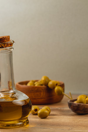 A glass bottle filled with golden olive oil sits on a wooden table next to wooden bowls filled with green olives, creating a rustic culinary setting ideal for cooking.の写真素材