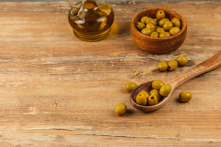 A collection of fresh green olives sits on a wooden spoon next to a small bowl filled with more olives. A bottle of olive oil is seen in the background, creating a rustic culinary display.の写真素材