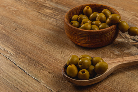 Fresh green olives are arranged in a wooden bowl and a spoon on a textured wooden table. The natural appearance enhances the rustic feel of the setting.の写真素材