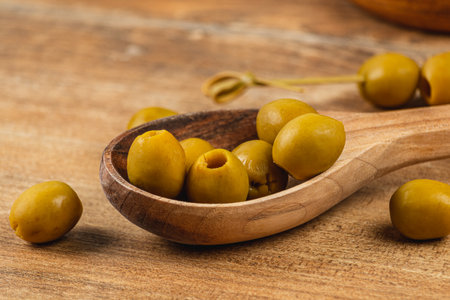 Fresh green olives are gathered in a wooden spoon, showcasing their natural shine and smooth texture. The rustic wooden table adds a warm touch to the arrangement of olives.の写真素材