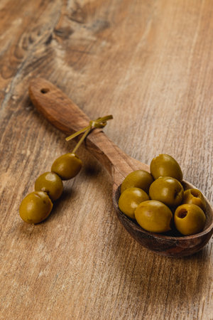 A wooden spoon holds several green olives, displaying their shiny texture. A few olives are scattered nearby on a rustic wooden table. The setting exudes a natural atmosphere.の写真素材