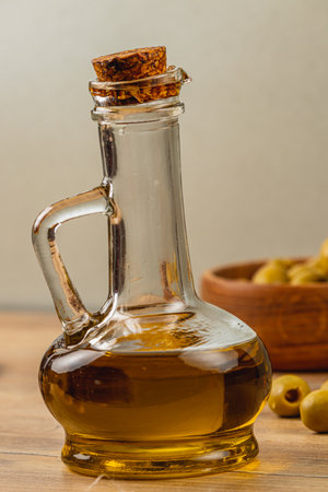 A glass bottle filled with olive oil stands on a wooden table, with a cork stopper. Nearby, a bowl contains ripe green olives, highlighting an Italian culinary theme.の写真素材