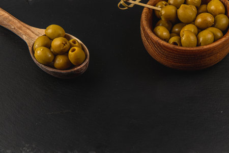 Delicious pitted green olives are arranged in a wooden bowl alongside a wooden spoon filled with olives, all set on a dark, textured surface.の写真素材