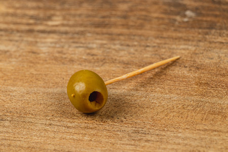 A green olive is skewered on a wooden toothpick and placed on a rustic wooden table. The simple presentation highlights the olive's rich color and smooth texture.の写真素材