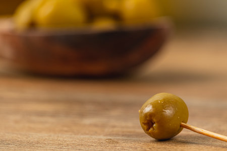 A single green olive rests on a toothpick on a wooden table. In the background, a bowl filled with more olives is slightly blurred, highlighting a rustic kitchen feel.の写真素材