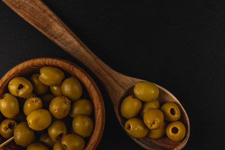 A wooden bowl filled with green olives sits alongside a wooden spoon, which also holds a few olives, set against a dark background, highlighting the natural textures.の写真素材