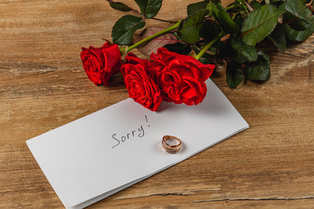 A heartfelt apology for a broken engagement is symbolized by a note saying Sorry, a wedding ring, and three red roses on a rustic wooden surface.の写真素材