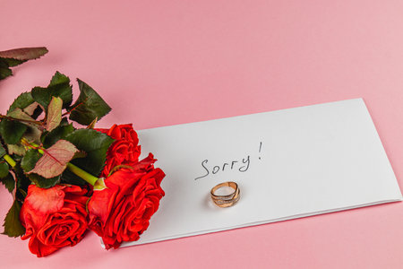 A pair of red roses rests beside a handwritten note saying Sorry and two intertwined wedding rings placed on a white envelope. This reflects a poignant moment of loss and farewell.の写真素材