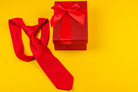 A bright yellow background highlights a red tie placed next to a gift box wrapped in vibrant red ribbon. Ideal for celebrating father's day or a birthday, this setup embodies thoughtful gifting.の写真素材