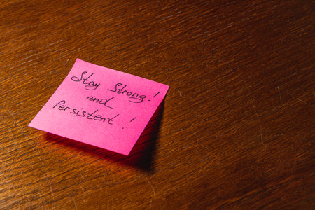 A pink sticky note with handwritten encouragement sits on a wooden table, conveying a motivational message to stay strong and persistent in challenging times.の写真素材