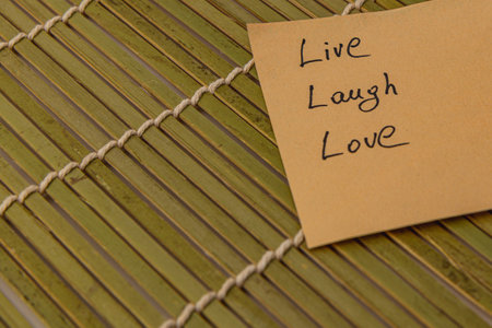 A note with the words live, laugh, love is placed on a textured bamboo mat. The simple message encourages positivity and mindfulness in a calm indoor setting.の写真素材
