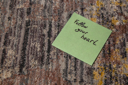 A green sticky note with the words follow your heart is laid on a multicolored carpet. The carpet features a mix of gray, brown, and yellow tones, creating a warm atmosphere.の写真素材