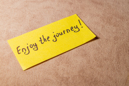 A bright yellow sticky note rests on a soft surface, featuring the handwritten message 'Enjoy the journey!' to inspire positivity and mindfulness in daily life.の写真素材