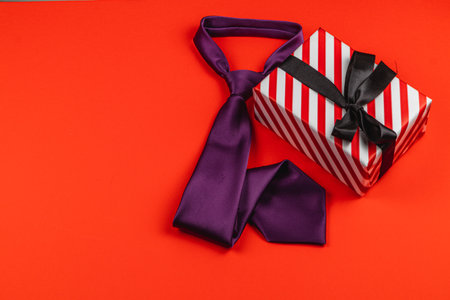 A stylish purple tie lies beside a neatly wrapped birthday gift on a vibrant red background. This arrangement symbolizes unique ways to honor fathers during celebrations.の写真素材