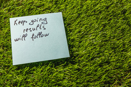 A light blue sticky note with handwritten motivational message is placed on vibrant green grass, reminding viewers to keep going as results will follow.の写真素材