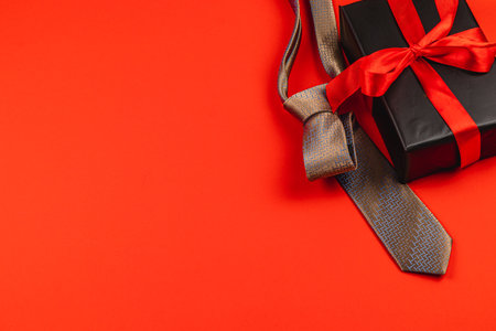 A beautifully wrapped gift box with a red ribbon sits alongside a patterned tie on a vibrant red surface.の写真素材
