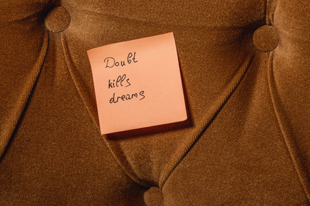 A sticky note with the message Doubt kills dreams is placed on a textured brown cushion, emphasizing the importance of self-belief and motivation, especially in personal growth.の写真素材