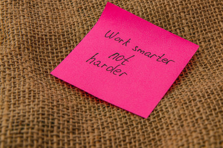 A bright pink sticky note rests on a rough burlap surface, displaying the motivational message about working efficiently instead of simply harder, encouraging productivity.の写真素材