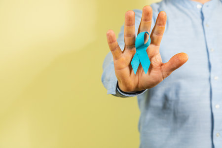 A man holds up a hand showing a blue ribbon, symbolizing prostate cancer awareness. He stands against a soft yellow background, representing support and hope for health issues affecting men.の写真素材