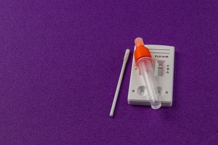 A flu testing kit is arranged on a vibrant purple background. The kit includes a test strip, a...