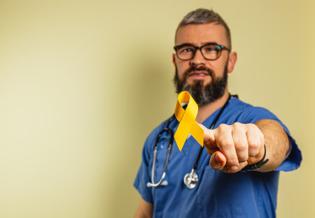 A young male healthcare worker with a stethoscope offers a yellow awareness ribbon in a clinic environment. He promotes health awareness with a smile, contributing to community support.の写真素材