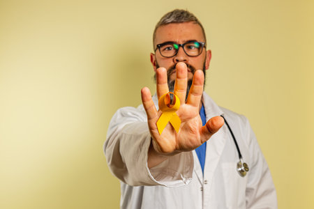 A young male healthcare worker displays a yellow ribbon, symbolizing awareness. He wears a white coat and stethoscope, smiling confidently in a well-lit clinic space.の写真素材