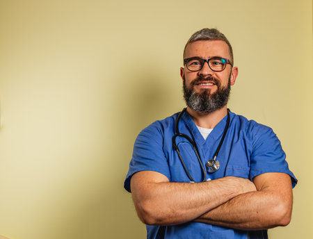 A young male doctor in blue scrubs stands confidently with arms crossed, smiling warmly in a clinic. He is prepared to assist patients in a supportive healthcare environment.の写真素材