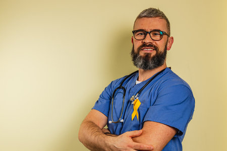 A happy male healthcare worker in blue scrubs stands with arms crossed, wearing a stethoscope and a yellow ribbon. He is smiling, embodying professionalism and care in a clinic setting.の写真素材