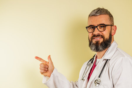 A young male doctor wearing glasses and a white coat is smiling and pointing to an empty space on the wall in a clinic setting, conveying a sense of welcome and positivity during a patient visit.の写真素材