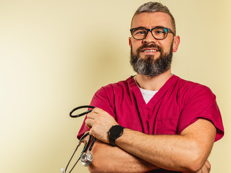 A young male cardiologist stands with his arms crossed, wearing scrubs and a stethoscope. He is in a clinic, exuding confidence and professionalism while smiling brightly.の写真素材