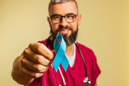 A young male cardiologist wearing scrubs and a stethoscope proudly displays a blue awareness ribbon. He smiles in a clinic environment, showing commitment to patient care and health advocacy.の写真素材