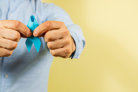 A person holds a blue awareness ribbon between their hands, symbolizing support for cancer prevention and awareness. The background is a soft yellow, emphasizing health and hope.の写真素材