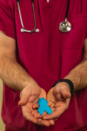 A male healthcare professional in scrubs holds a blue awareness ribbon in his hands, smiling as he promotes health awareness in a clinic space. He wears a stethoscope around his neck.の写真素材