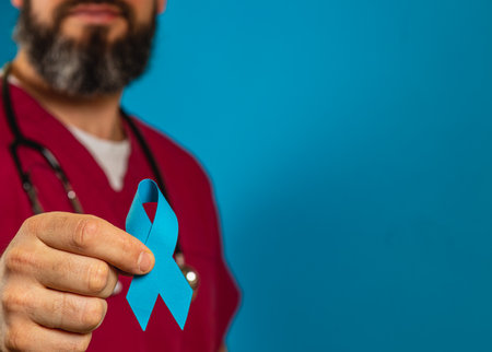 A young, smiling male doctor in scrubs holds a blue awareness ribbon in a clinic. He displays a sense of support and care, emphasizing the importance of health awareness in patient care.の写真素材