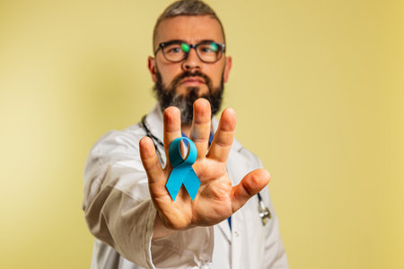 A male doctor in scrubs and glasses is smiling while holding a blue awareness ribbon in front of him. He stands in a clinical setting, promoting health and support for illness awareness.の写真素材