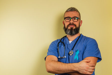 A happy male nurse in blue scrubs stands with arms crossed in a clinic. He wears glasses and a stethoscope, exemplifying professionalism and dedication to patient care.の写真素材
