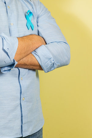 A man stands with crossed arms wearing a light blue shirt, proudly displaying a blue ribbon symbolizing awareness for cancer on International Cancer Day, set against a soft yellow background.の写真素材