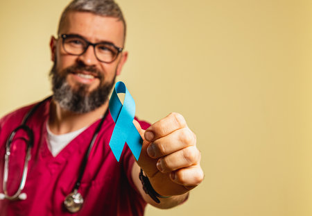 A male cardiologist in burgundy scrubs and glasses smiles while holding a blue awareness ribbon in a clinic. He promotes health and care for patients with illness or disease.の写真素材