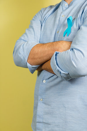 A person with crossed arms wearing a light blue shirt adorned with a blue ribbon symbolizing prostate cancer awareness stands against a yellow background, promoting health awareness.の写真素材