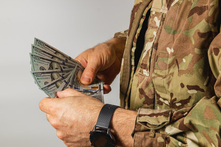 A soldier in a camouflage uniform is holding a stack of cash, counting the bills attentively while...