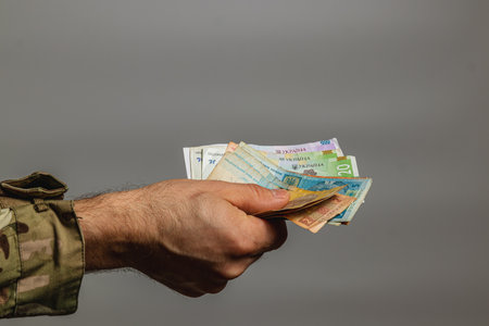 A hand holds various currency notes against a gray background. The person's attire suggests a military influence, adding a unique context to the exchange.の写真素材
